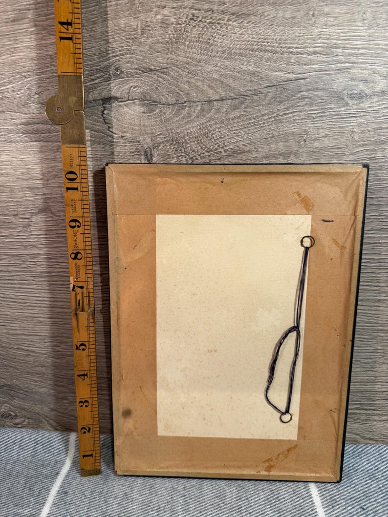 May include: The back of a framed picture with a brown paper backing. A vintage folding ruler is next to the frame. The frame is rectangular and has a black border. The ruler is made of wood and brass and measures up to 14 inches.