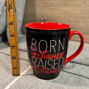 Peut inclure: Mug en c&eacute;ramique noire avec int&eacute;rieur et anse rouges. Le mug porte l'inscription "BORN DISNEY RAISED DISNEY" en lettres blanches et rouges. Le mug mesure environ 10 cm de haut.