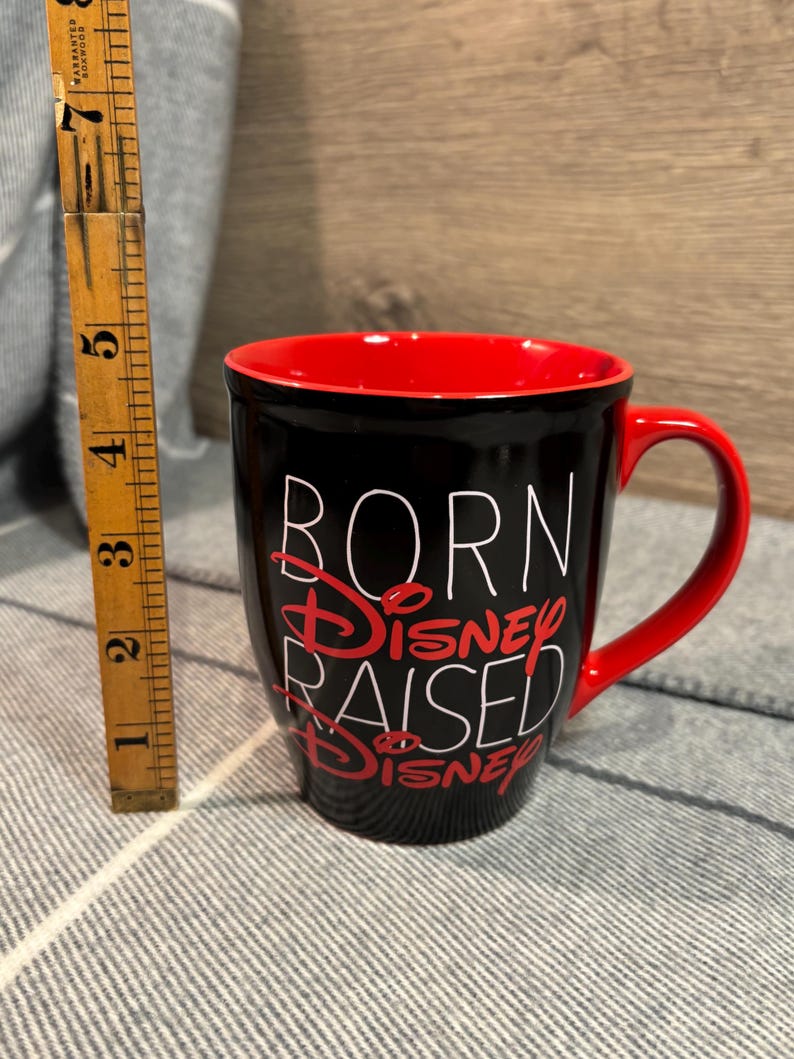 Peut inclure: Mug en c&eacute;ramique noire avec int&eacute;rieur et anse rouges. Le mug porte l'inscription "BORN DISNEY RAISED DISNEY" en lettres blanches et rouges. Le mug mesure environ 10 cm de haut.