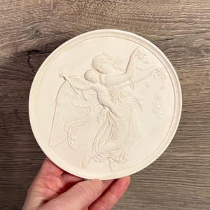 Op de afbeelding: Een ronde, crèmekleurige reliëfsculptuur met een klassieke scène van een gevleugelde figuur die een kind vasthoudt. De figuren dragen vloeiende gewaden, met fijne details. De sculptuur wordt in een hand gehouden.