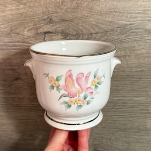 May include: White ceramic planter with a gold rim and base. The planter features a floral design with pink and yellow flowers and green leaves. It has two small handles on the sides.