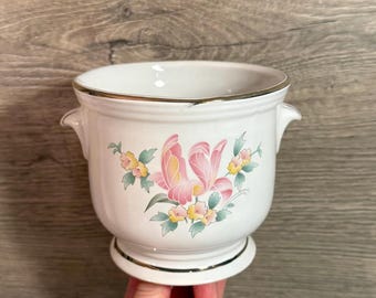 May include: White ceramic planter with a gold rim and base. The planter features a floral design with pink and yellow flowers and green leaves. It has two small handles on the sides.