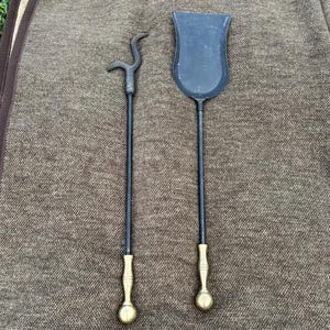 Op de afbeelding: Een haardgereedschapsset met een pook en een schop. De gereedschappen hebben zwarte schachten met messingkleurige handvatten en punten. De pook heeft een gebogen uiteinde en de schop heeft een afgeronde schep.