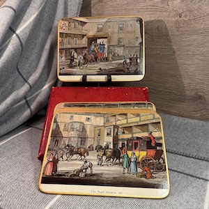 May include: Set of vintage coasters featuring historical illustrations of buildings and horse-drawn carriages. The coasters have a rectangular shape with a gold trim and are stored in a red box. The text "The Angel" and "The Queen's Head" are visible.