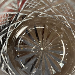 Op de afbeelding: Close-up van een helder glas met ingewikkelde geslepen kristallen patronen. Het ontwerp heeft geometrische vormen en radiale lijnen die uit het midden komen en een sterrenburst-effect cre&euml;ren. Het glas is waarschijnlijk voor dranken.