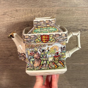 May include: A ceramic teapot with a castle-like design, depicting Robin Hood and his companions in Sherwood Forest. The teapot has a white handle and spout with gold detailing. The text reads "Robin Hood in Sherwood Forest."
