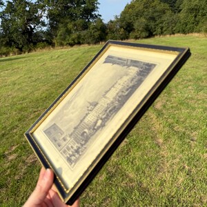 May include: A framed black and gold print of a building with a detailed architectural design. The print is held at an angle against a backdrop of green grass and trees under a blue sky.