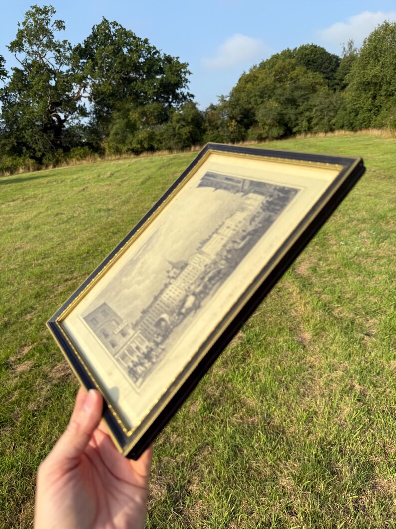May include: A framed black and gold print of a building with a detailed architectural design. The print is held at an angle against a backdrop of green grass and trees under a blue sky.