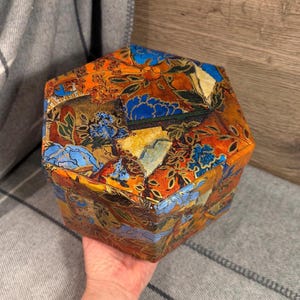 May include: A hexagonal box featuring a vibrant, multi-coloured collage pattern with shades of orange, blue, yellow, and black. The surface displays intricate floral and abstract motifs across the lid and sides, creating a detailed, textured appearance.