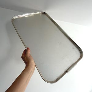 Puede incluir: Una bandeja de servicio rectangular de metal plateado con esquinas redondeadas y una superficie texturizada. La bandeja cuenta con un borde metálico liso y elevado, además de dos asas integradas en los lados más cortos.