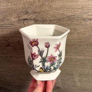 May include: White ceramic vase with a hexagonal top and base. The vase is decorated with pink and purple flowers, green leaves, and a yellow butterfly. The text "Anemone Pulsatilla" is printed on the side.