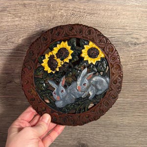 May include: A round cast iron trivet with a detailed relief design. The trivet features two grey rabbits nestled among green leaves and yellow sunflowers. The border is a dark brown with an ornate, floral pattern.