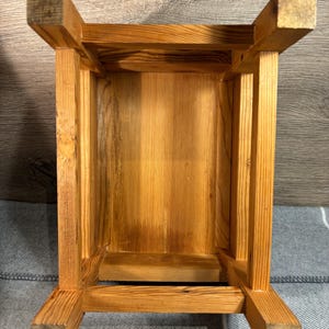 May include: A wooden stool, viewed from underneath, showcasing its construction. The stool is made of light-colored wood with visible grain. The design features a rectangular seat supported by four square legs, joined by horizontal supports.