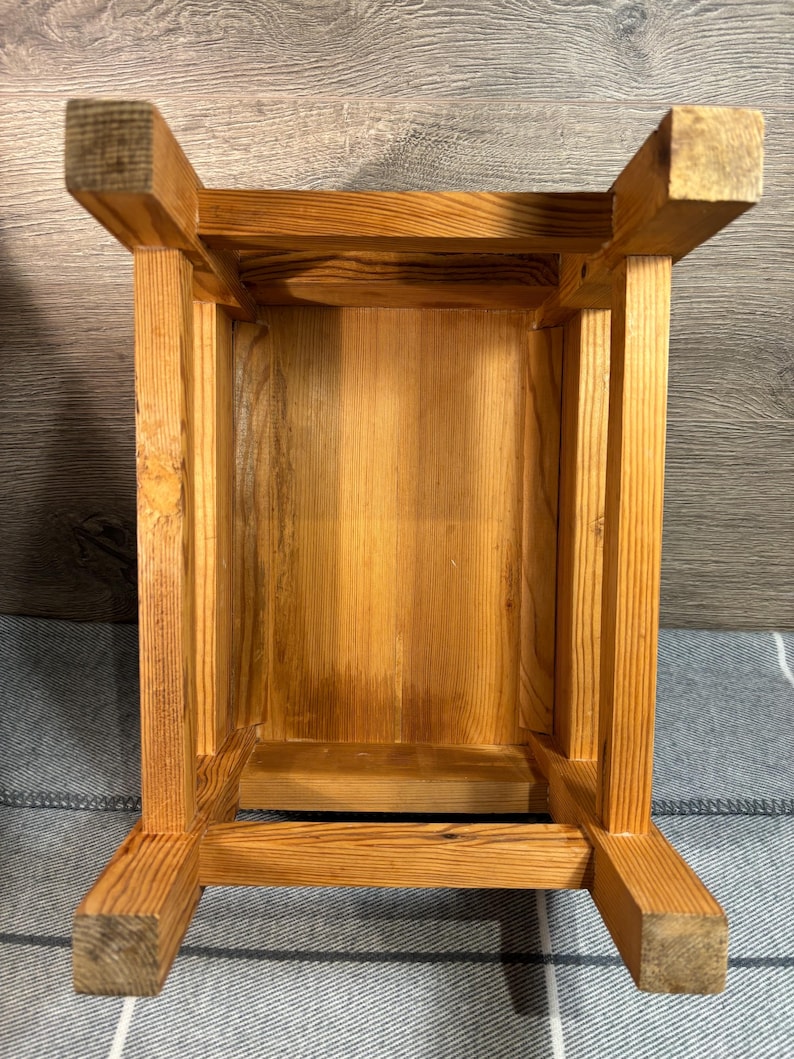 May include: A wooden stool, viewed from underneath, showcasing its construction. The stool is made of light-colored wood with visible grain. The design features a rectangular seat supported by four square legs, joined by horizontal supports.