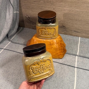 May include: Two ceramic kitchen canisters, one labelled "Coffee" and the other "Tea". The canisters are tan with brown lids and decorative lettering. The "Tea" canister sits atop a wooden stand. The "Coffee" canister is held by a hand.