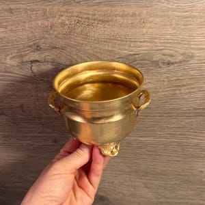 Puede incluir: Un cuenco pequeño y redondo de metal dorado con un acabado liso en el interior y el exterior. El cuenco presenta dos asas decorativas y ornamentadas en los lados opuestos y se apoya sobre tres pies pequeños y detallados en la base.