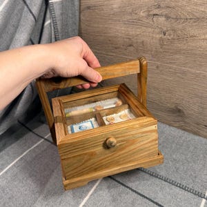 Könnte beinhalten: Eine Holz-Nähkiste mit Griff, Schublade und durchsichtigem Deckel. Die Kiste enthält Garnrollen, Knöpfe und eine kleine Briefmarke. Die Kiste ist aus hellbraunem Holz.