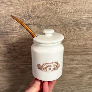 Pode incluir: Pote de cerâmica branca com tampa e colher de pau. O pote apresenta uma ilustração castanha de uma casa. O pote é segurado por uma pessoa. O pote é provavelmente para guardar condimentos ou outros artigos de cozinha.