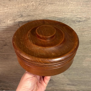 May include: A round wooden bowl with a lid. The bowl is made of dark brown wood and has a smooth finish. The lid is also made of wood and has a knob on top.