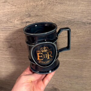May include: A dark blue ceramic mug featuring a gold emblem with a crown, the text 1952 1977, the letters EIIR, and the words SILVER JUBILEE. The mug has a cylindrical body with a raised circular medallion on the front and a square-shaped handle.