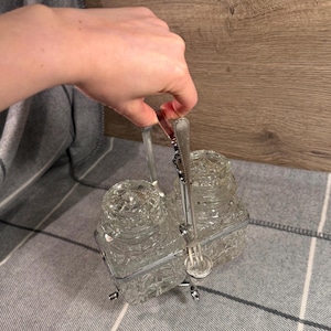 Pode incluir: Um conjunto vintage de saleiro e pimenteiro de vidro com um suporte de metal prateado. Os saleiros de vidro transparente têm um design de vidro lapidado texturizado. Inclui um garfo pequeno. O conjunto é segurado por uma pessoa.