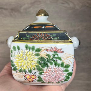 Op de afbeelding: Een witte keramische container met een deksel, versierd met gele, roze en rode bloemmotieven. Het deksel en de randen zijn afgewerkt met goud en zwart. Het heeft twee kleine witte handvatten.
