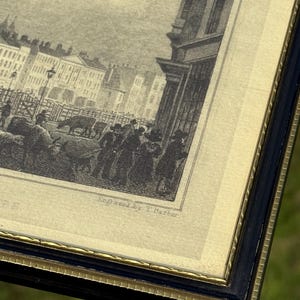 May include: An antique framed print with a black and gold frame. The print is a black and white engraving of a street scene with buildings, people, and animals. The text "Engraved by T. Barber" is visible.