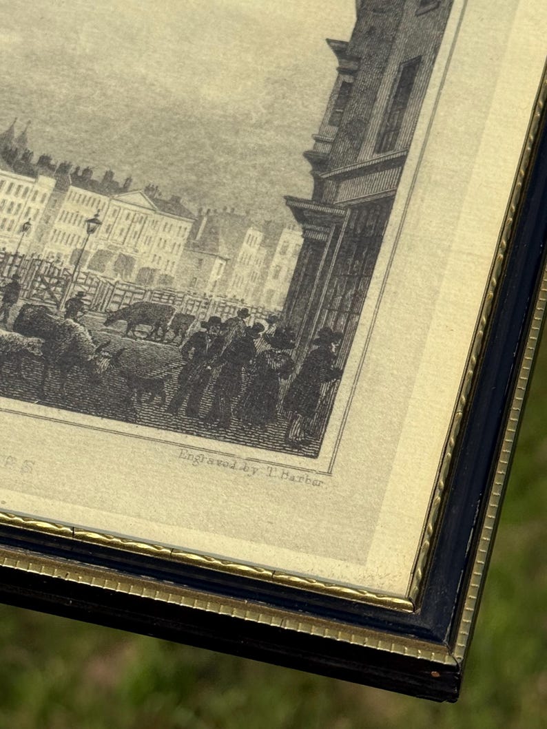 May include: An antique framed print with a black and gold frame. The print is a black and white engraving of a street scene with buildings, people, and animals. The text "Engraved by T. Barber" is visible.