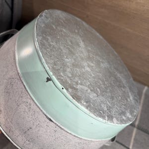 Op de afbeelding: Een ronde metalen emmer met een afwerking van gegalvaniseerd staal aan de bovenkant en de basis, geaccentueerd door een brede band van licht muntgroene verf in het midden. De container heeft twee kleine metalen handvatten aan de zijkanten.