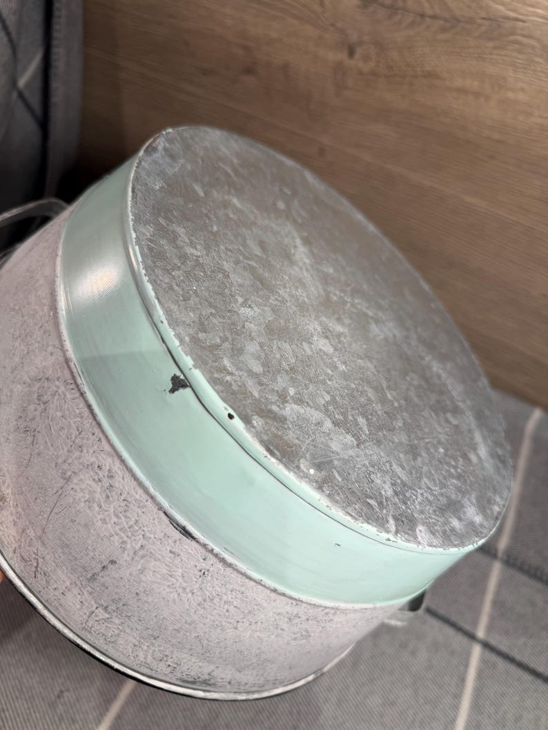 Op de afbeelding: Een ronde metalen emmer met een afwerking van gegalvaniseerd staal aan de bovenkant en de basis, geaccentueerd door een brede band van licht muntgroene verf in het midden. De container heeft twee kleine metalen handvatten aan de zijkanten.