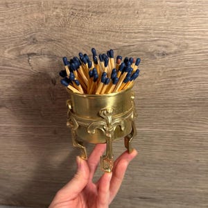 May include: A brass-colored match holder filled with matches. The holder has an ornate design with three legs and a round bowl. The matches have blue tips and are arranged upright in the holder.