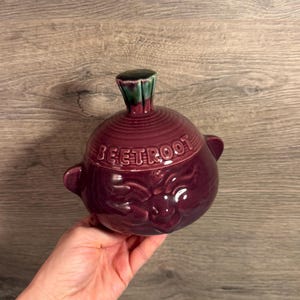 May include: A deep purple ceramic storage jar with a lid, featuring the word BEETROOT embossed on the side. The jar has two small side handles and a green-tipped knob on the lid. The surface is glossy and smooth with a rounded, bulbous shape.