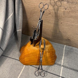 Puede incluir: Dos pinzas de servir plateadas, una sobre un soporte de madera marrón oscuro y la otra sobre una superficie texturizada marrón claro. Las pinzas tienen un diseño similar a unas tijeras con mangos redondeados. El fondo incluye una tela estampada gris y blanca y una pared de madera.