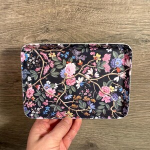 Peut inclure: Plateau rectangulaire décoratif avec un motif floral. Le plateau présente un fond noir avec des fleurs roses, bleues, jaunes et blanches et des feuilles vertes. Le plateau est tenu par une main et repose sur une surface en bois.