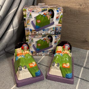May include: A quirky butter dish set with a ceramic design of two figures in a bed, covered with a green blanket. The dish has a purple base and a dark brown headboard. The box has the words "BUTTER DISH".