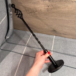 Könnte beinhalten: Ein schwarzer Metall-Kaminhaken mit einem dekorativen Spiraldesign am Schaft und einem kreisförmigen Sockel. Die Oberseite hat ein dekoratives Wirbeldesign und eine kleine Kugel. Der Haken wird von einer Person gehalten.