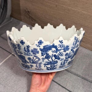 Pode incluir: Uma tigela de cerâmica branca com borda recortada, apresentando um padrão azul sob o vidrado de peixes koi, plantas aquáticas e flores. A superfície externa é decorada com ilustrações detalhadas de peixes a nadar e elementos botânicos.