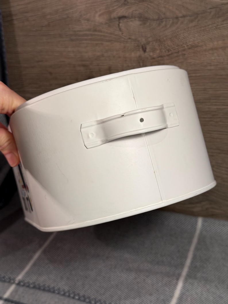 May include: A cylindrical white metal storage container featuring a horizontal handle attached to the side. The container has a smooth, solid white finish with a visible vertical seam running down the side and a small central hole in the handle.