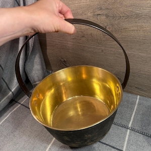 May include: A vintage metal cauldron with a dark exterior and a bright gold interior. The cauldron has a rounded shape and a sturdy, arched handle. The handle is made of a darker metal, contrasting with the gold interior. The cauldron is held by a hand.