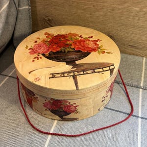May include: A round decorative box with a floral design. The lid features a painting of a vase filled with red and pink flowers on a table. The box has a red cord handle and a floral design around the sides.