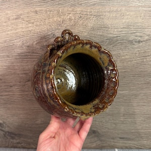 Op de afbeelding: Een handgemaakte keramische vaas met een getextureerde, gevlekte bruine en bruingele glazuur. De vaas heeft een brede opening en een decoratieve, geschulpte rand. De bovenkant van de vaas heeft drie kleine, geluste handvatten. De binnenkant is donker, glanzend bruin.