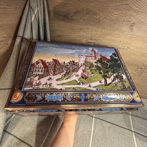 May include: A rectangular, vintage metal box with a detailed, colorful illustration of a European townscape. The box features a blue and gold decorative border and a hinged lid. The image includes text.