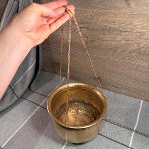 May include: A round, metallic brass bowl with a weathered, aged patina finish. The bowl features a wide, open top and a rounded base. It is suspended by three thin, gold-toned metal chains that meet at a single point for hanging display.