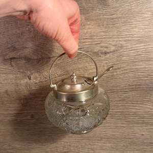 Peut inclure: Un sucrier en verre transparent avec un couvercle et une poignée argentés. Le bol présente un motif décoratif texturé. Une petite cuillère repose sur le couvercle. La poignée est tenue par une main.
