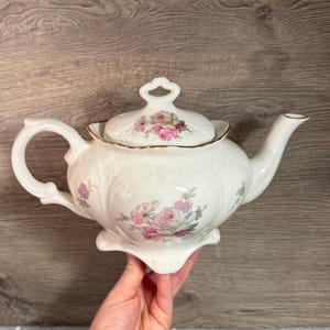 May include: White porcelain teapot with a floral rose design in pink and green. The teapot has a curved spout, a loop handle, and a lid with a matching floral pattern. The edges are trimmed with gold.