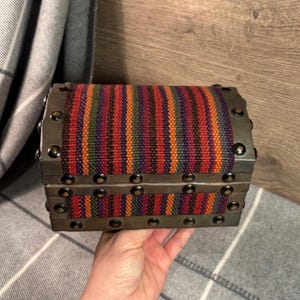Op de afbeelding: Een kleine houten schatkist met een kleurrijk gestreept stoffen deksel en metalen accenten. De kist is versierd met metalen studs rond de randen.
