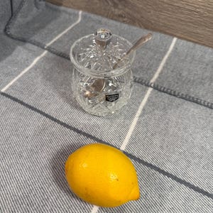 Op de afbeelding: Een heldere kristallen suikerpot met een deksel en een kleine lepel erin. Een heldergele citroen ligt naast de kom op een grijs en wit gestreept oppervlak. De kom heeft een decoratief geslepen ontwerp.