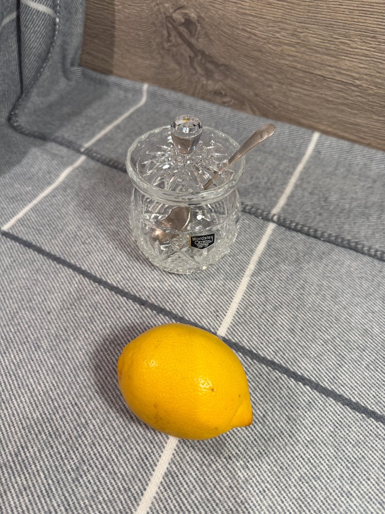 Op de afbeelding: Een heldere kristallen suikerpot met een deksel en een kleine lepel erin. Een heldergele citroen ligt naast de kom op een grijs en wit gestreept oppervlak. De kom heeft een decoratief geslepen ontwerp.