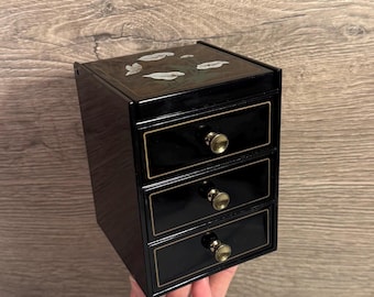 Op de afbeelding: Een zwarte sieradendoos met drie lades en goudkleurige knoppen. De bovenkant van de doos heeft een decoratief ontwerp van witte bladeren. De doos is rechthoekig en heeft een glanzende afwerking.