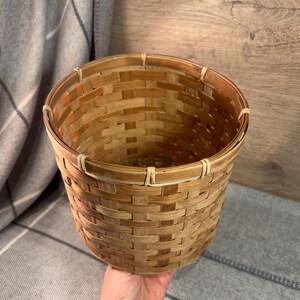 May include: A woven brown bamboo planter with a natural finish. The planter is round and has a wide opening. It is made of natural materials and has a rustic look.